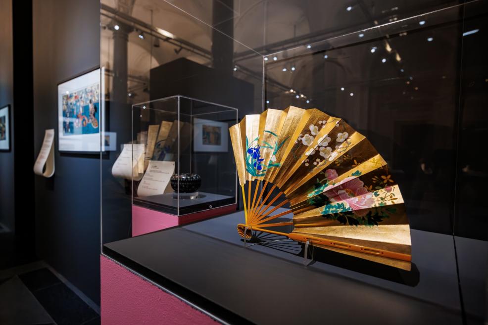 A golden Japanese folding fan with floral motifs displayed in a museum case, with other exhibits and prints visible in the background.