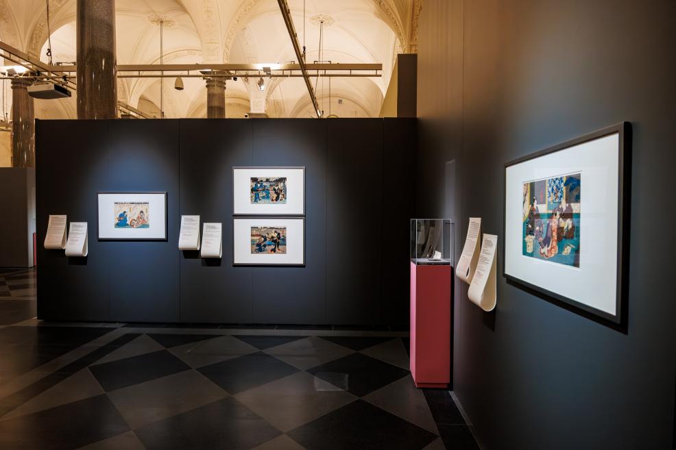 A museum exhibition with dark walls displaying framed Japanese prints and explanatory texts, with columns and vaulted ceilings visible in the space.