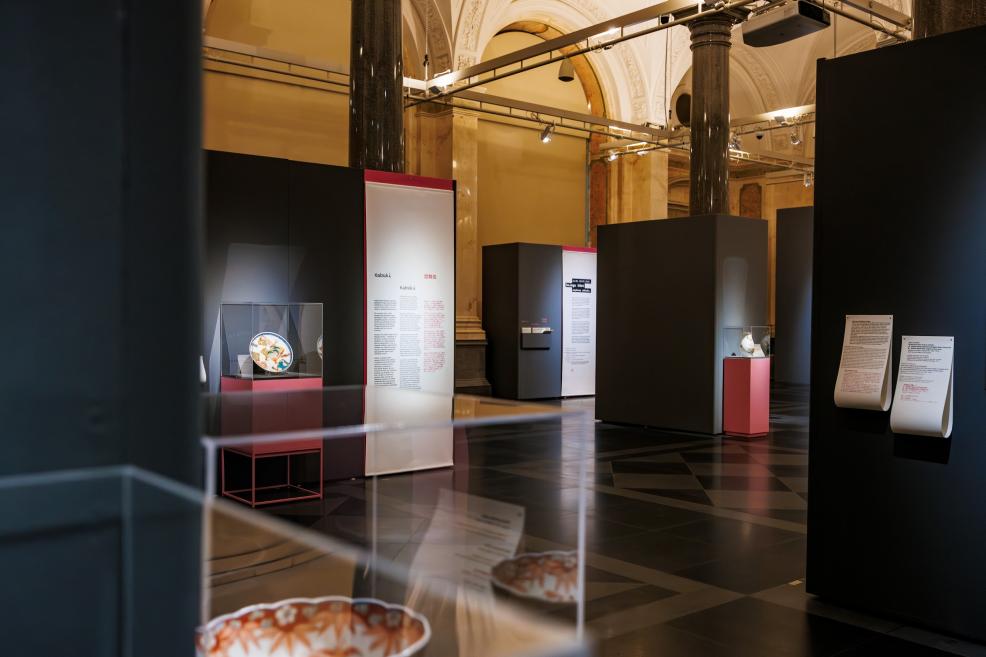 A view of a museum exhibition with dark display structures, glass cases containing ceramic objects, and informational panels, with columns and vaulted ceilings in the background.