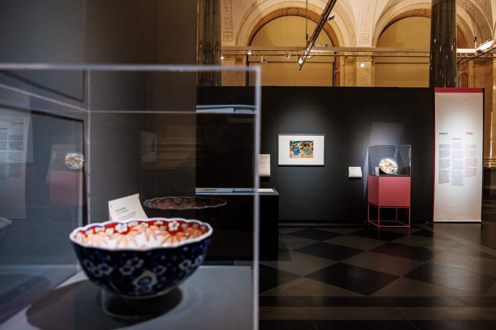 A view of a museum exhibition featuring a decorative ceramic bowl displayed in a glass case, with Japanese prints and an informational panel visible in the background.