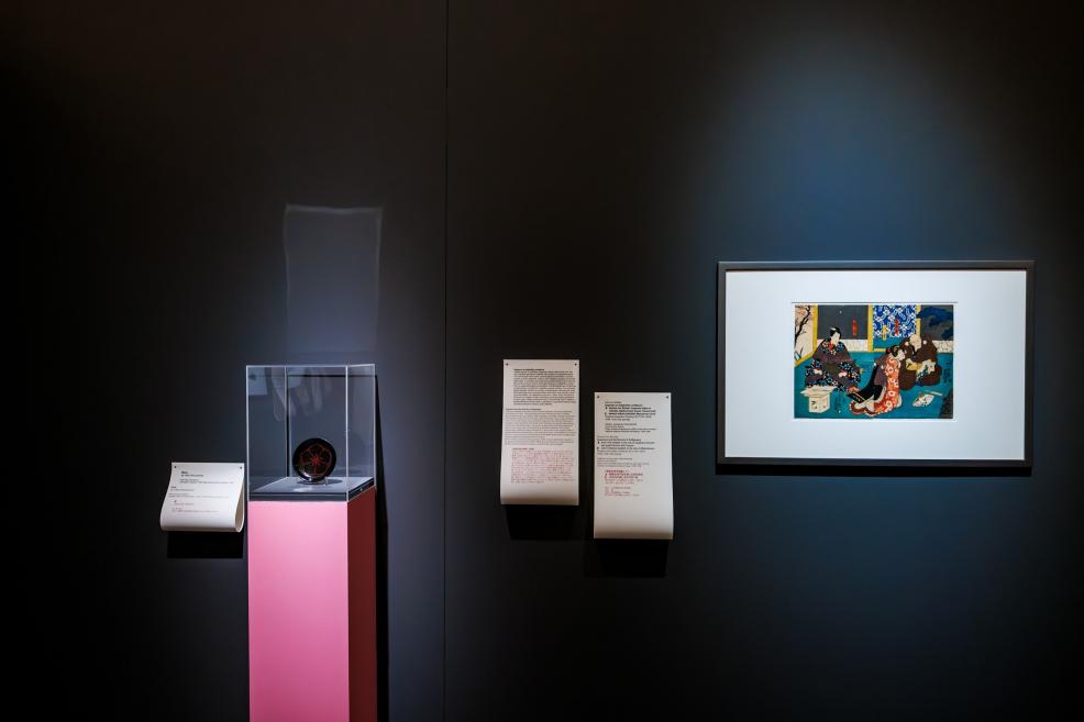 A section of a museum exhibition featuring a small decorative object displayed in a glass case on a pink pedestal, accompanied by informational texts and a framed Japanese color print on the wall.