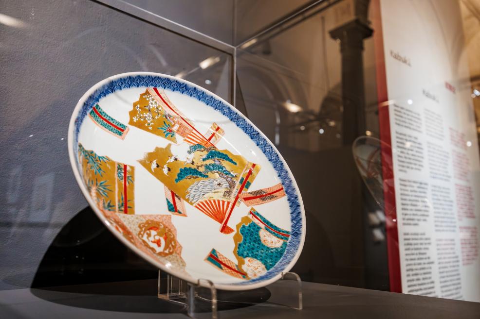A decorative porcelain plate with a colorful Japanese motif displayed in a glass case, with an informational panel visible in the background.