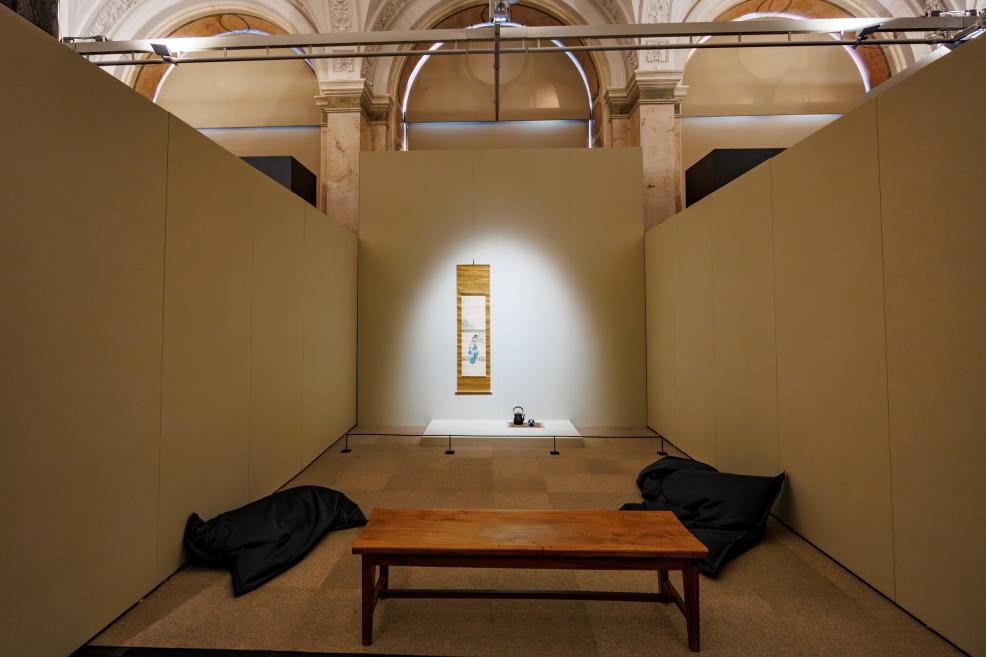 A museum exhibition space featuring a centrally illuminated vertical Japanese hanging scroll, with a low table and floor cushions for visitors.