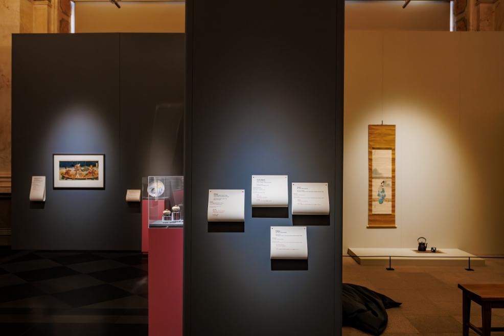 Muzeja ekspozīcijas skats ar sienām izvietotām japāņu grafikām, stikla vitrīnu uz rozā postamenta un blakus telpā izgaismotu vertikālu gleznojuma rulli.