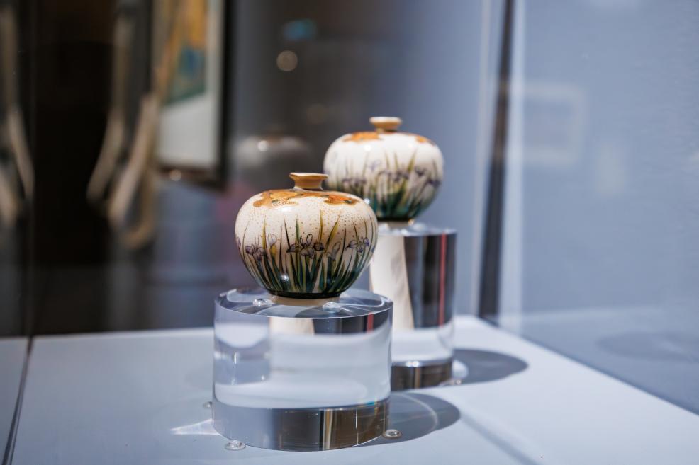Two small decorative ceramic vessels with floral motifs displayed in a glass case on transparent stands.