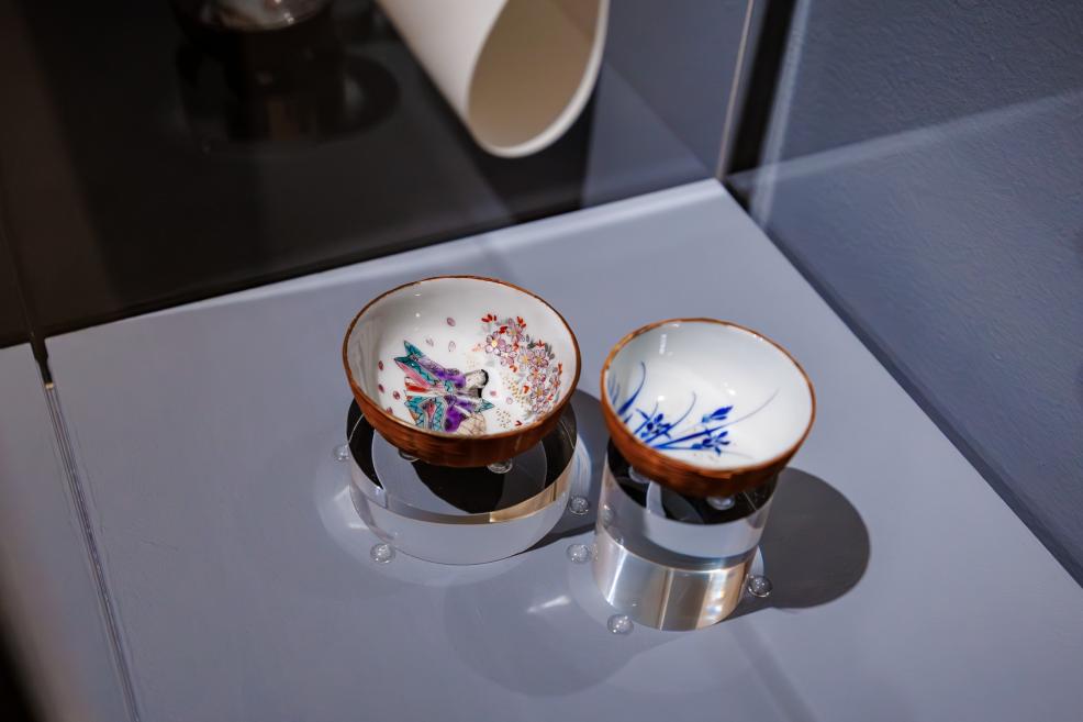 Two small porcelain bowls with colorful floral and figure motifs displayed in a glass case on transparent stands.