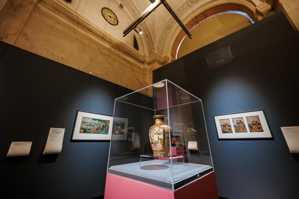 A decorative vase displayed in a glass case on a pink pedestal within a museum exhibition, with framed Japanese prints and historic hall architecture visible in the background.