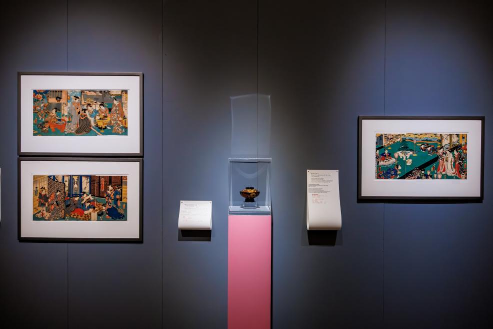 A museum exhibition wall featuring several framed Japanese color prints and a glass case on a pink pedestal displaying a small decorative vessel.