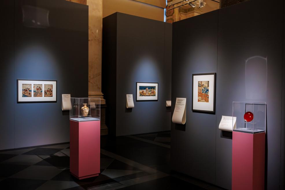 A museum exhibition space with framed Japanese prints on dark walls and glass display cases on pink pedestals presenting decorative objects.