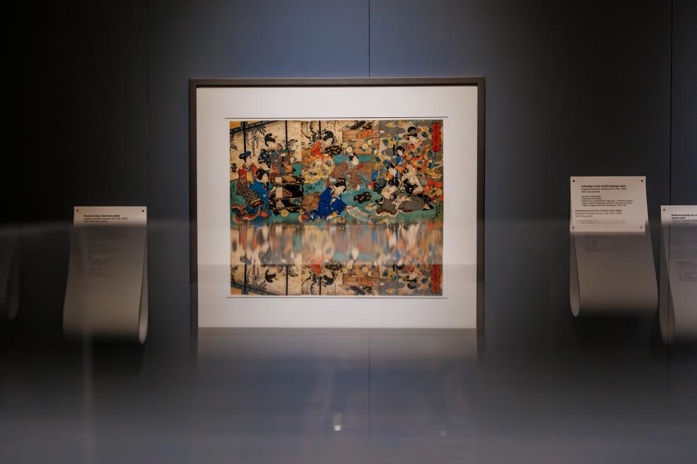 A colorful framed Japanese print depicting multiple figures indoors is displayed on a dark museum wall, with its lower part reflected in a glass surface and explanatory labels placed on both sides.