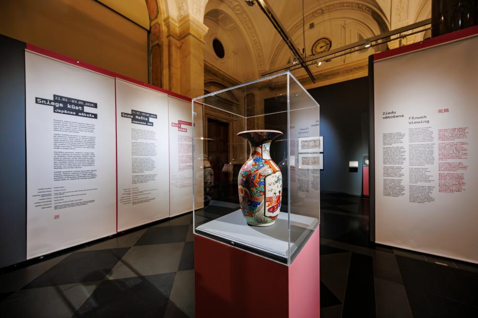 In a museum exhibition, a decorative Japanese vase is displayed in a glass case on a red pedestal, with large informational panels featuring exhibition texts visible in the background.