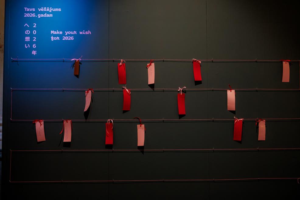 Red and white wish tags are attached to stretched strings on a dark wall, with the text “Tavs vēlējums 2026. gadam / Make your wish for 2026” visible in the upper left corner.