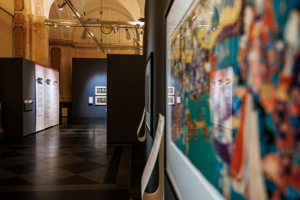 A museum exhibition hall with dark display panels and informational stands, with a blurred framed Japanese print visible in the foreground.