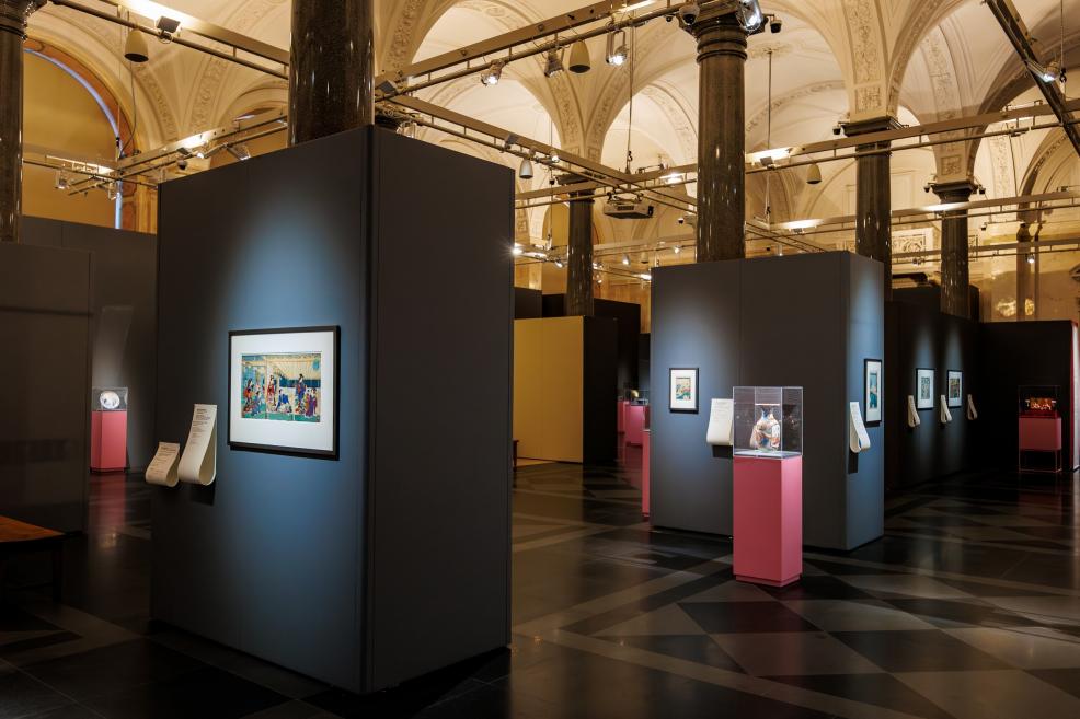 A spacious museum exhibition hall with high vaulted ceilings, dark display panels, framed Japanese prints, and glass cases on pink pedestals.