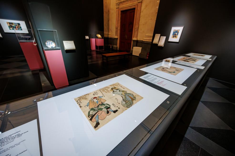 Japanese prints with captions are displayed horizontally in a museum showcase, with dark exhibition panels, glass cases on pink pedestals, and a bench visible in the background.