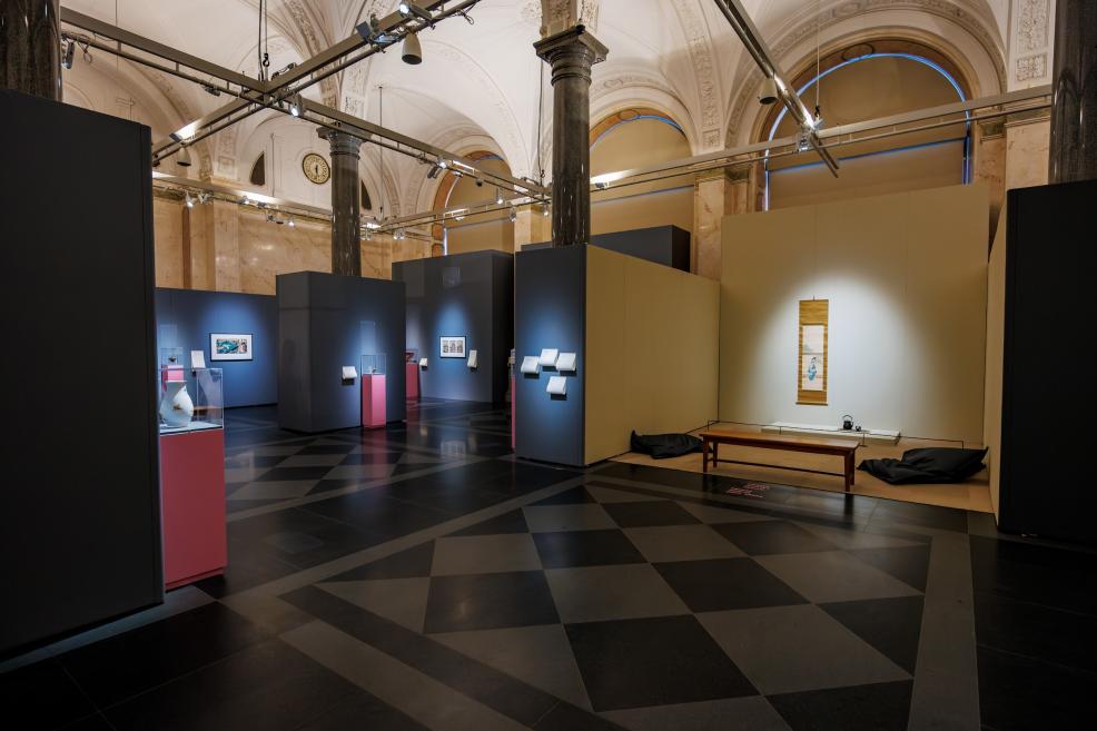 A spacious museum hall with high vaulted ceilings, dark exhibition panels, and glass cases on pink pedestals, with a calm seating area on the right featuring a hanging scroll and a bench.
