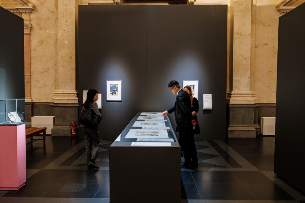 Three visitors are viewing Japanese prints displayed in a museum showcase, standing in front of a dark exhibition panel with framed works and labels in the background.
