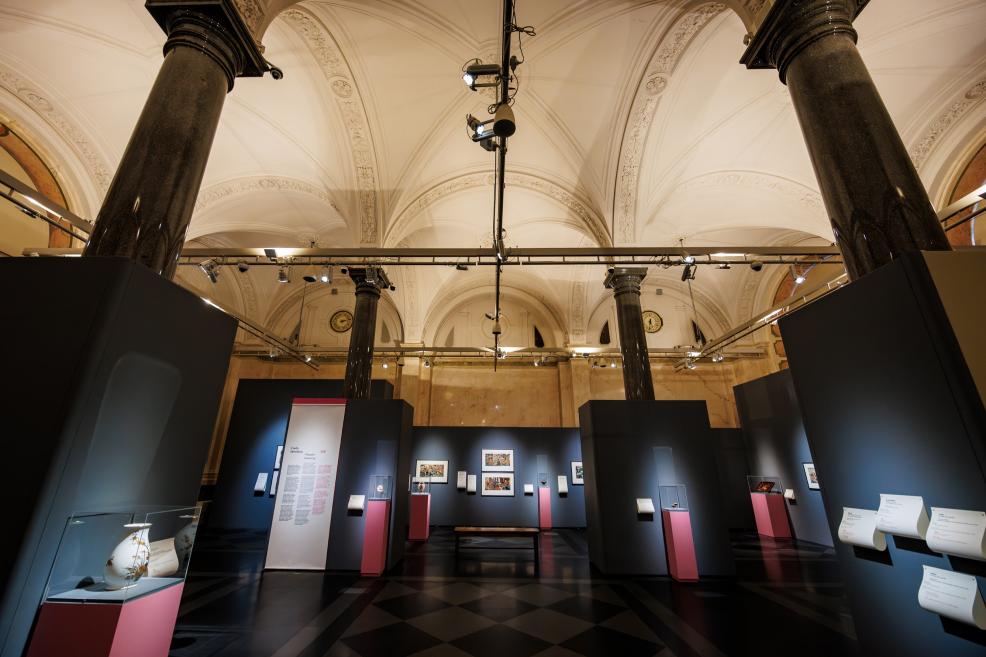 A museum exhibition hall with high vaulted ceilings and columns, featuring Japanese prints displayed on dark panels and glass cases on pink pedestals.