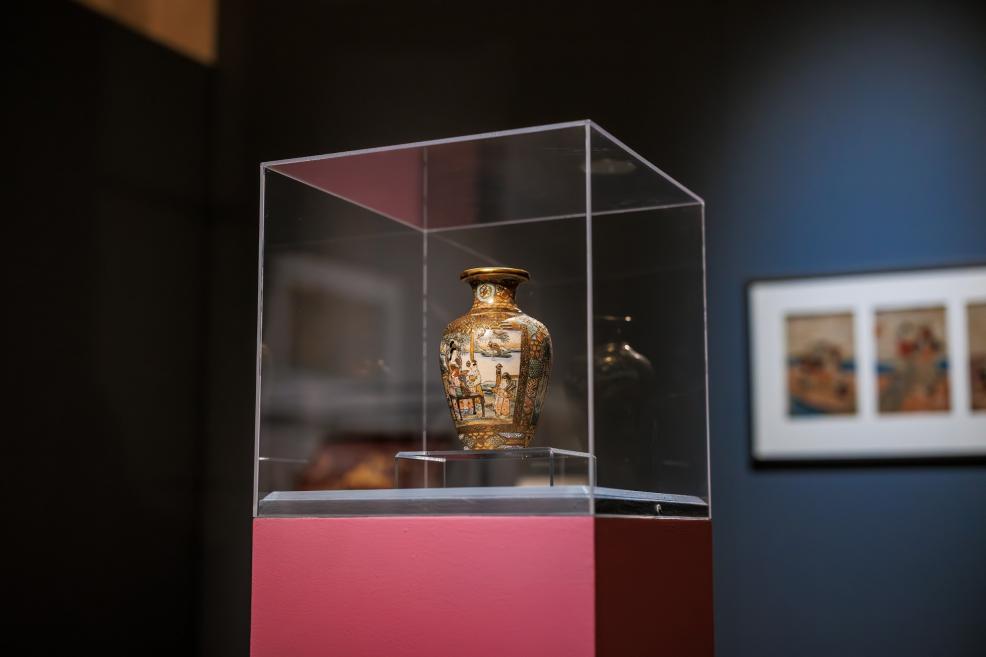 A small decorative Japanese vase with figurative scenes is displayed in a glass case on a pink pedestal against a dark exhibition panel.
