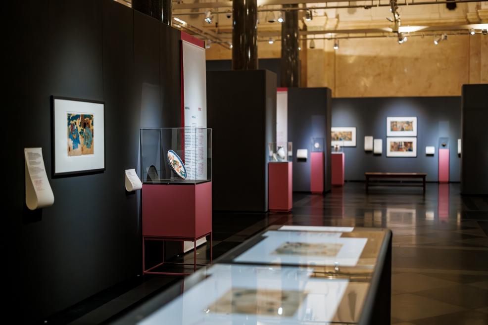 View of the exhibition hall with Japanese prints on dark panels, display cases on pink pedestals, and a glass display table with artworks in the foreground.