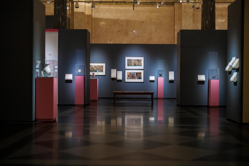 Symmetrical view of the exhibition hall with Japanese prints on dark panels, glass display cases on pink pedestals, and a bench in the center.