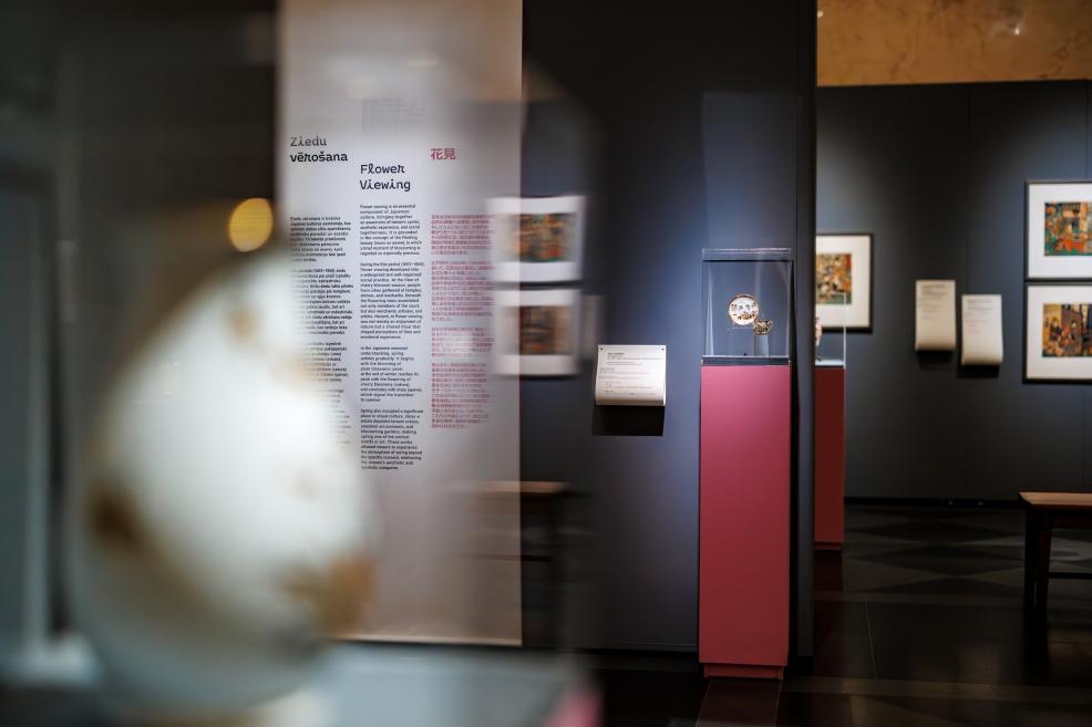 View of the exhibition section with the wall text “Ziedu vērošana / Flower Viewing” and a small decorative object displayed in a glass case on a pink pedestal.