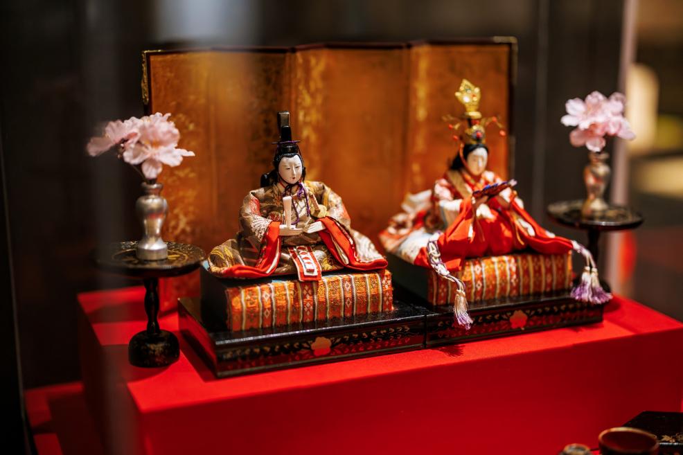 A pair of traditional Japanese figures in ceremonial attire displayed on a red pedestal with a decorative backdrop and flowers in vases.