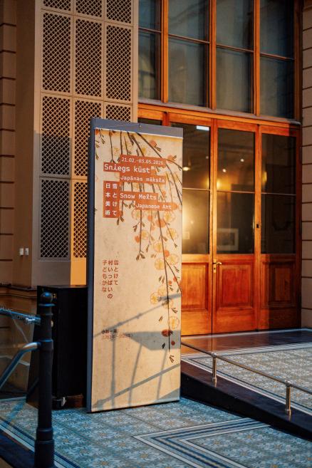 Exhibition sign reading “Sniegs kūst. Japānas māksla / Snow Melts. Japanese Art” at the museum entrance with wooden doors in the background.