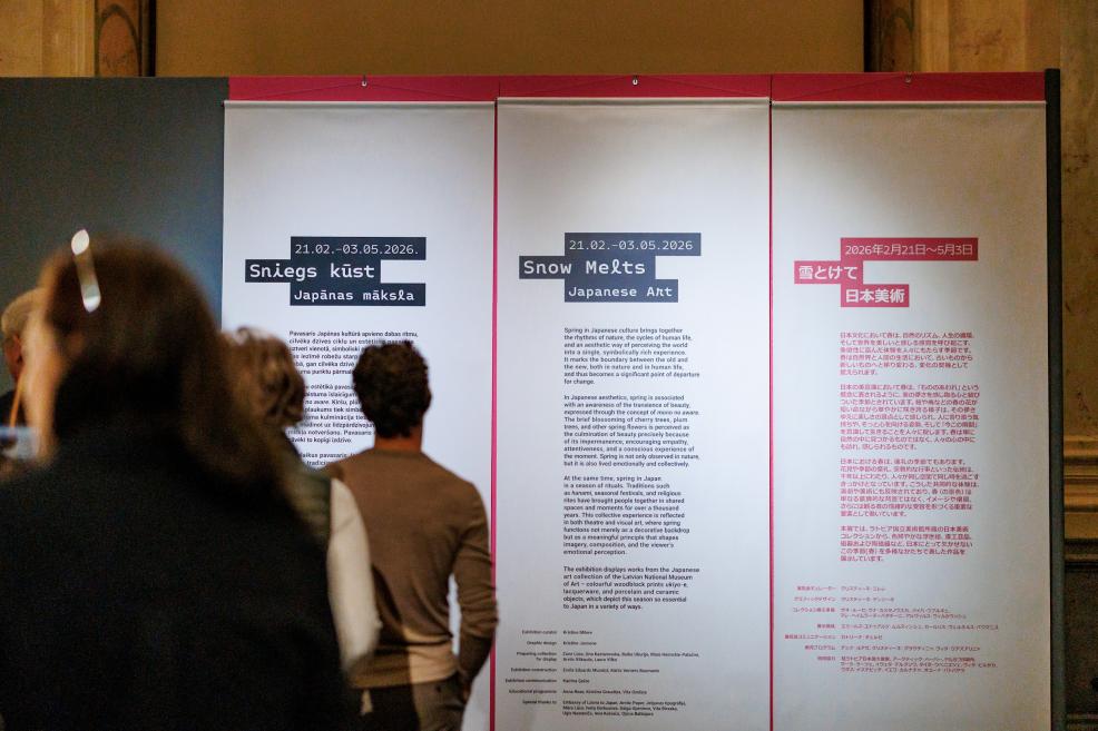 Visitors reading the exhibition introduction “Sniegs kūst. Japānas māksla / Snow Melts. Japanese Art” displayed on large wall panels.