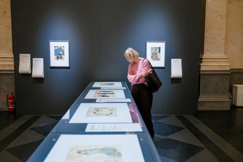 A visitor examining Japanese prints in a glass-topped display table, with framed works visible on a dark panel in the background.