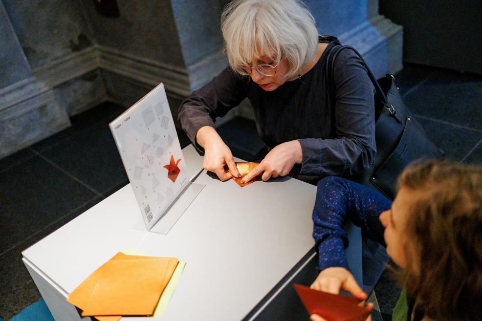 Sieviete un bērns pie galda saloka oranžu papīru origami tehnikā, izmantojot instrukciju kartīti.