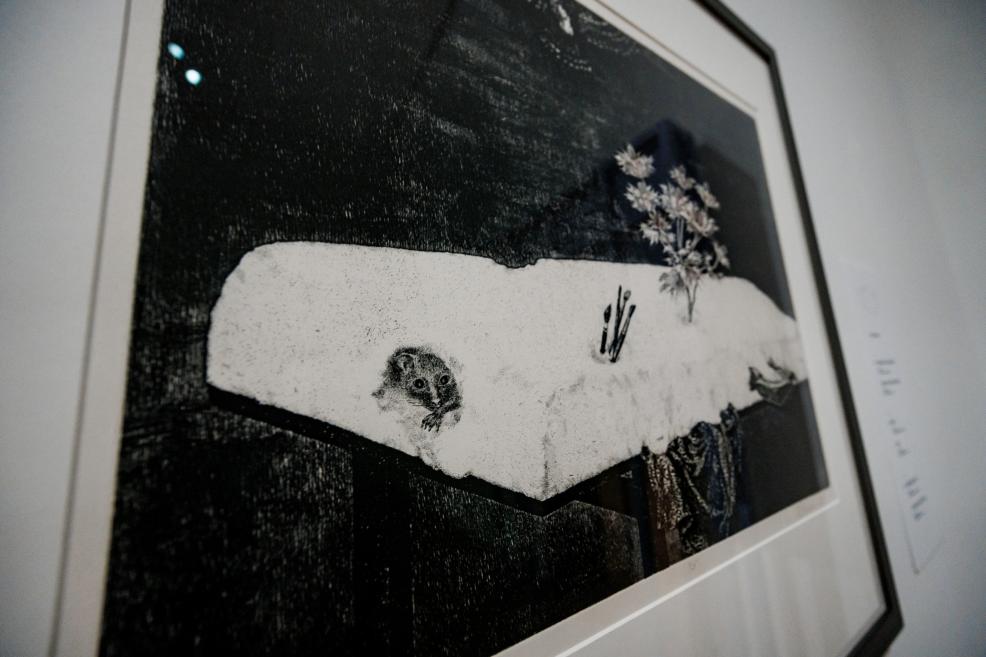 A framed black-and-white graphic artwork depicting a stylized landscape with a small animal and a plant motif set against a dark background.