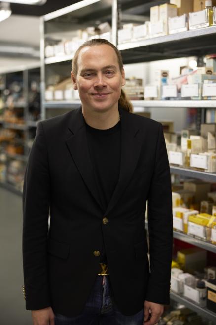 Color photograph of a man in a black jacket standing in the Roche archive among shelves of documents and packaging.