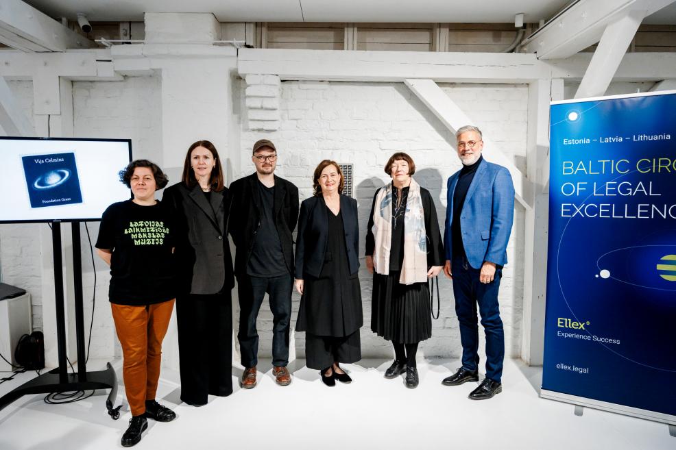 Seši cilvēki stāv izstāžu telpā pie Vijas Celmiņas fonda stipendijas prezentācijas stenda un informatīva reklāmas banera.