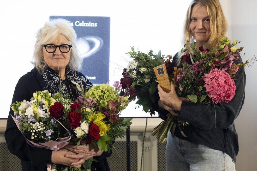 Divas sievietes tur lielus ziedu pušķus un Vijas Celmiņas fonda stipendijas balvu, stāvot pasākuma telpā pie prezentācijas ekrāna.