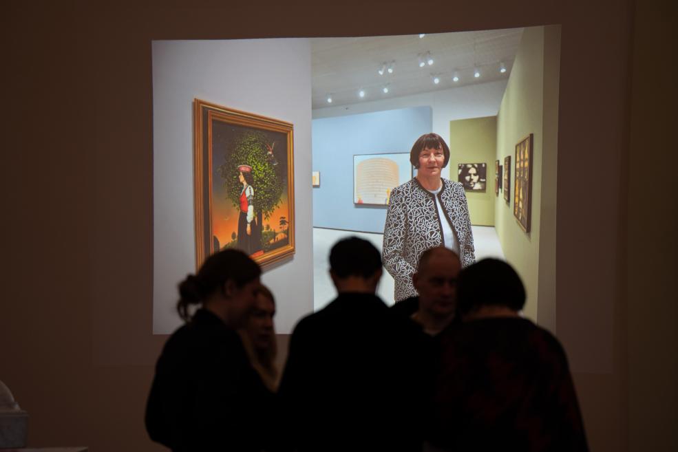 Uz sienas projicēts video ar sievieti mākslas galerijā, priekšplānā redzami cilvēku silueti.
