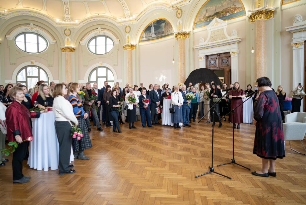 Cilvēku grupa ar ziediem klausās runu greznā muzeja zālē ar kolonnām un augstiem logiem.