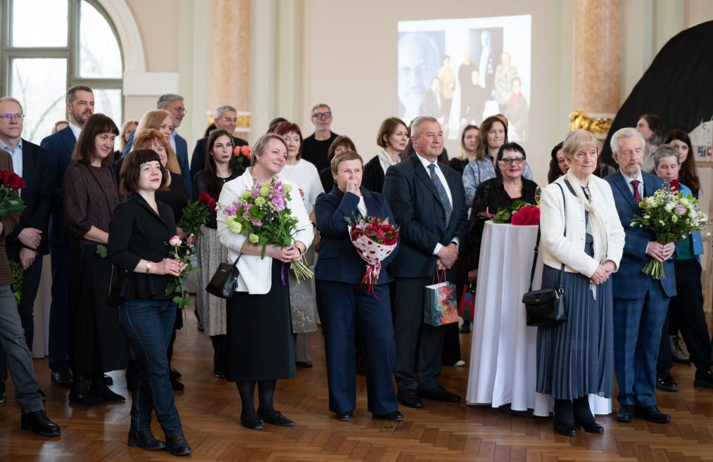 Dažāda vecuma cilvēki ar ziedu pušķiem stāv un klausās pasākumu muzeja zālē.