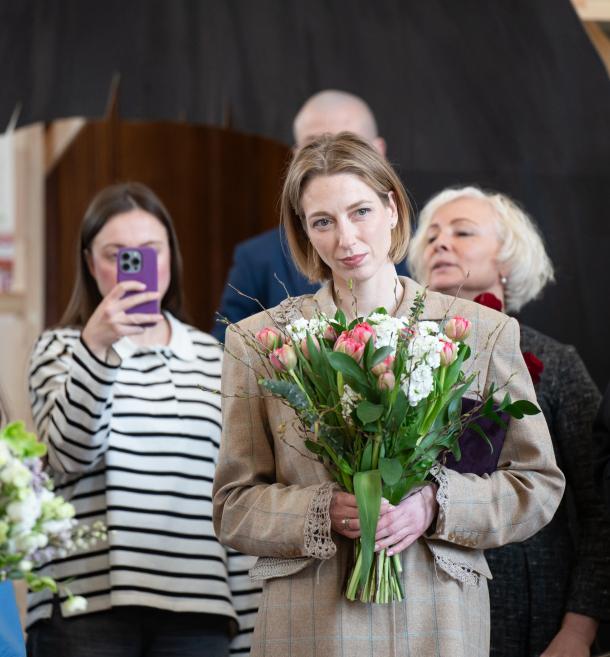 Sieviete ar tulpju pušķi rokās klausās pasākumu, kamēr fonā cita persona fotografē ar telefonu.