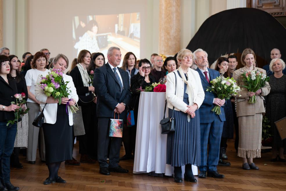 Cilvēki ar ziedu pušķiem stāv un klausās pasākumu muzeja zālē, fonā redzama projekcija.