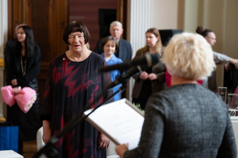 Sieviete stāv auditorijas priekšā, kamēr cita persona pie mikrofona lasa dokumentu muzeja pasākumā.