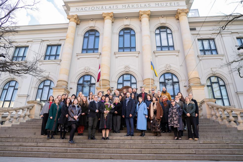 Liela cilvēku grupa stāv uz Latvijas Nacionālā mākslas muzeja kāpnēm pie ēkas ar kolonnām un karogiem.