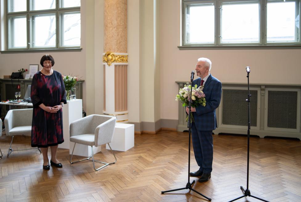 Vīrietis ar ziedu pušķi runā pie mikrofona muzeja zālē, kamēr sieviete stāv malā un klausās.