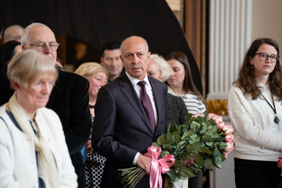 Vīrietis uzvalkā ar rožu pušķi stāv starp citiem cilvēkiem un klausās muzeja pasākumā.