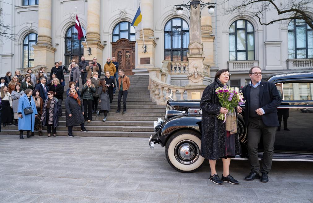 Sieviete ar ziedu pušķi un vīrietis stāv pie melnas retro automašīnas, kamēr cilvēku grupa kāpj lejup pa muzeja kāpnēm aiz viņiem.