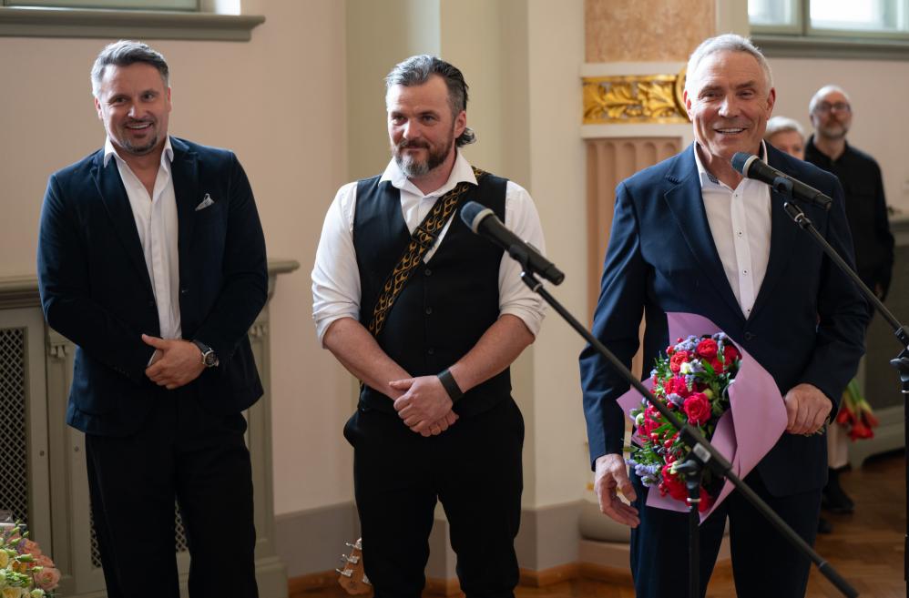 Trīs vīrieši stāv pie mikrofoniem muzeja zālē, viens no viņiem tur rokās ziedu pušķi.