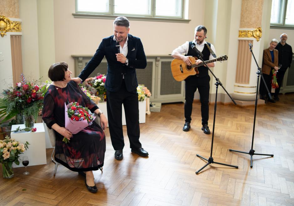 Vīrietis ar mikrofonu uzrunā sēdošu sievieti ar ziedu pušķi, kamēr blakus stāv mūziķis ar ģitāru muzeja zālē.
