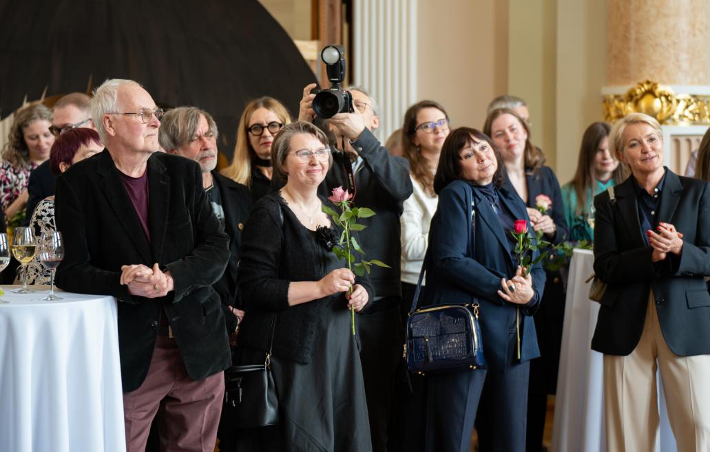 Cilvēku grupa muzeja zālē klausās un aplaudē, vairākiem rokās ir rozes, bet viens fotografē ar kameru.