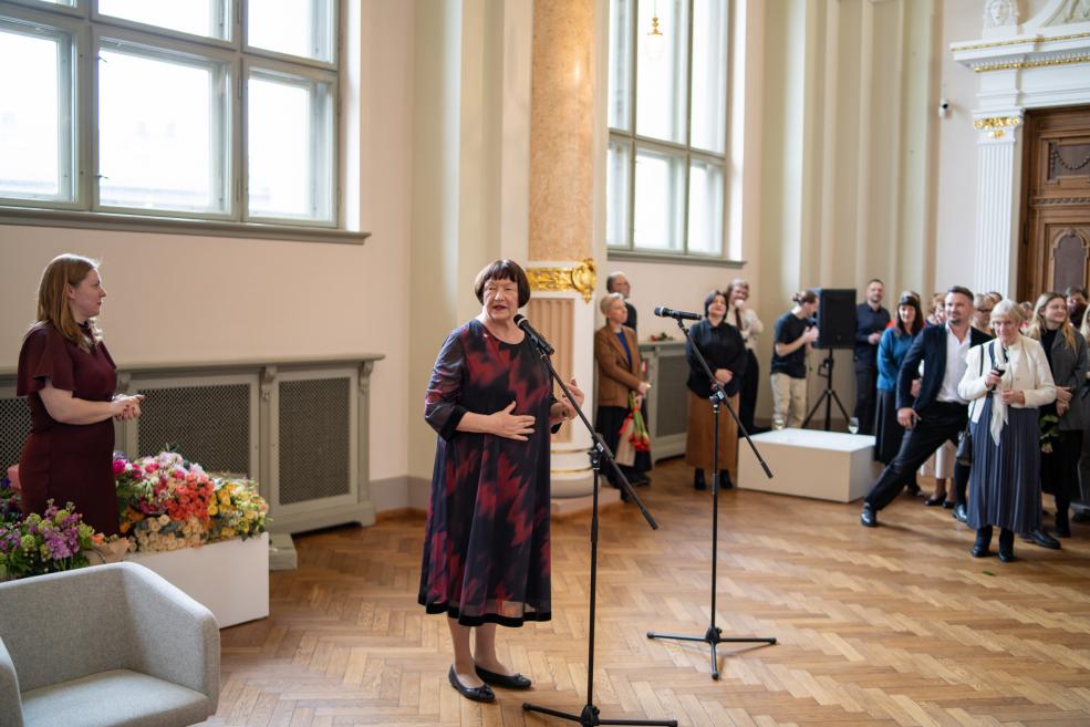 Sieviete stāv pie mikrofona un uzrunā klātesošos muzeja zālē, kamēr apkārt pulcējušies cilvēki klausās.