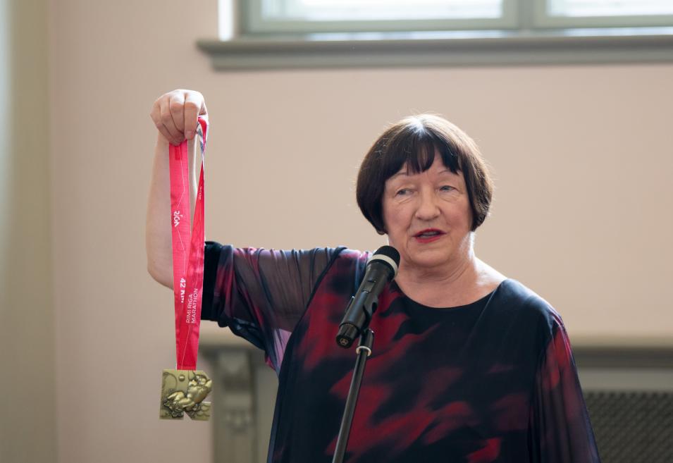 Sieviete pie mikrofona paceļ rokā medaļu ar sarkanu lenti un runā muzeja zālē.